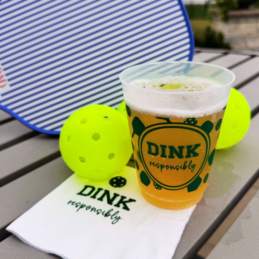 A Fresh Pickle Pickleball Roadie Cup labeled “DINK responsibly” sits on a matching napkin beside two yellow pickleballs and a blue-striped paddle on a gray outdoor table, with greenery and a blurred patio in the background.