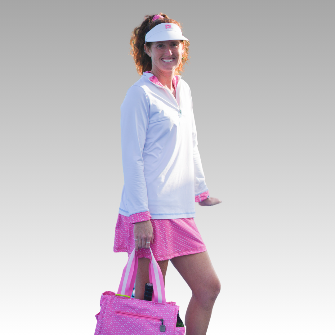 A smiling woman stands against a gray background wearing the Playly Store Soleil Tech Shirt, a white long sleeve made from breathable fabric. She pairs it with a pink skirt, white visor, and holds a pink-patterned tote. Soft natural lighting highlights her look.