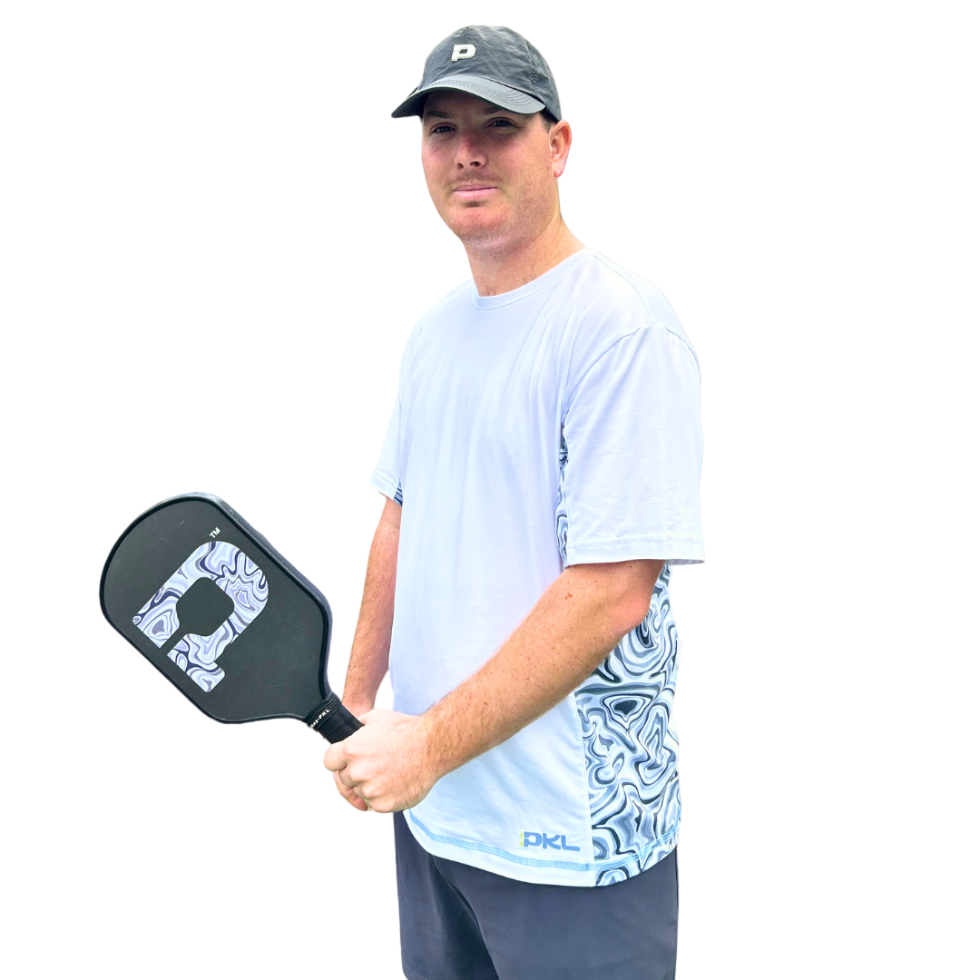 A man in a light blue patterned Playly Store Warrior Tee 2.0, black shorts, and a black cap holds a black pickleball paddle, standing against a simple gray gradient background and facing slightly to the right while looking at the camera.