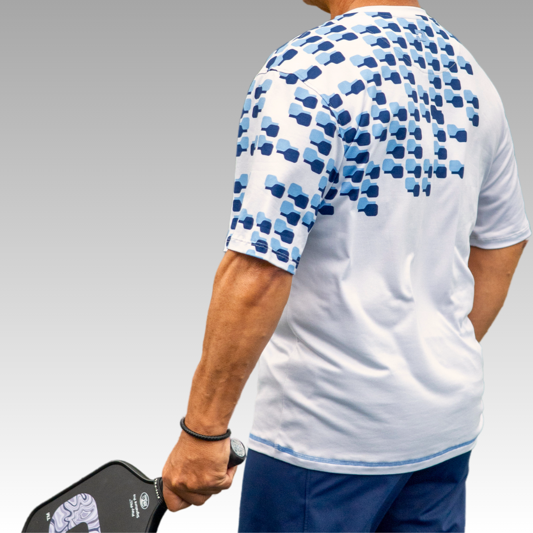 A person stands facing away, holding a pickleball paddle in their right hand. They wear the Playly Store Playly - Warrior Tee 2.0, a white performance shirt with a blue geometric pattern on the upper back, paired with blue shorts against a gray-to-white background.