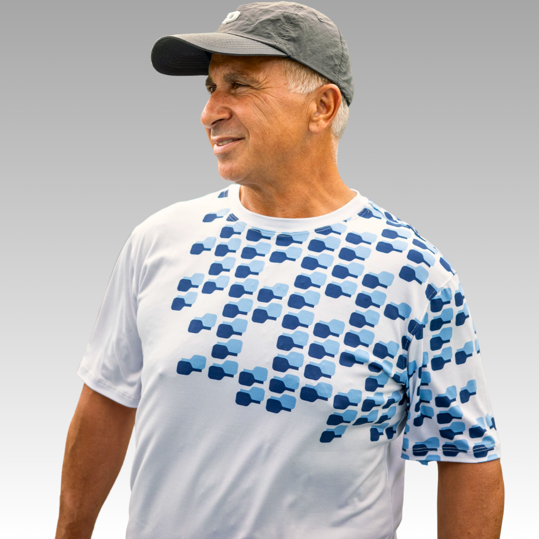 An older man in a gray cap wears the Playly Store’s Playly - Warrior Tee 2.0, a moisture-wicking t-shirt with rows of dark and light blue shapes, as he smiles and looks left against a plain gray background.