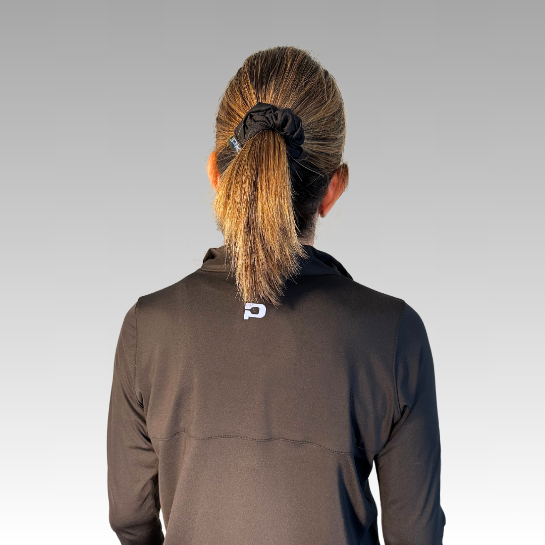 A person with light brown hair in a ponytail, secured by a Shanti Pickleball Hair Scrunchie from Playly Store, wears a black jacket with a white P logo on the back and stands against a gray gradient background.