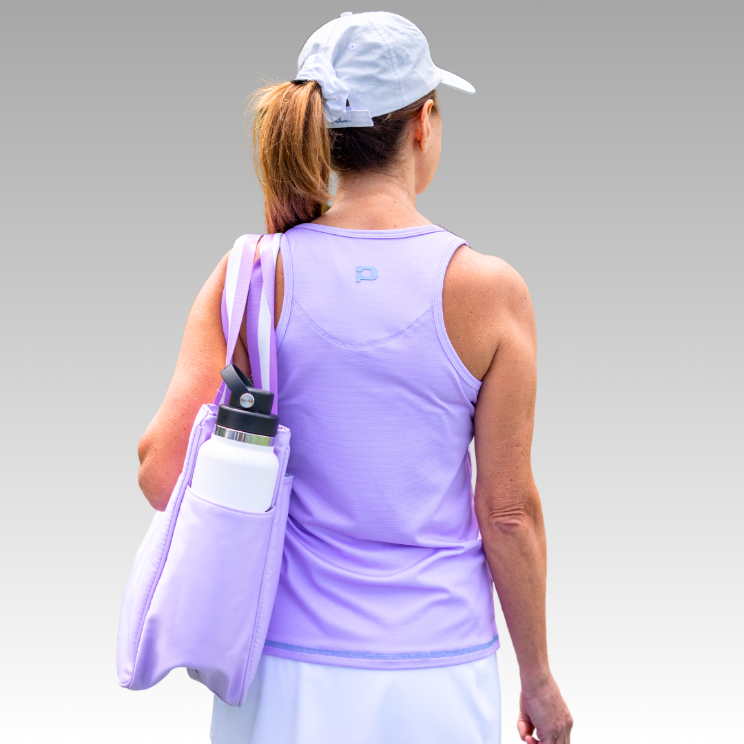 A woman with light skin and brown ponytail wears the Playly Store Pretty Tank in light purple, paired with a white cap and skirt. She carries a matching purple tote bag and white bottle, standing against a gradient gray background.