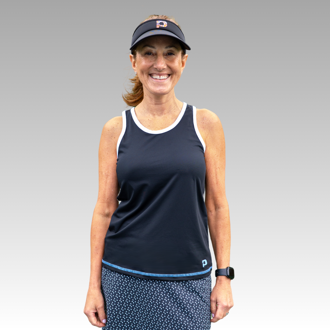 A smiling woman with reddish-brown hair wears the Playly Store Pretty Tank, a black moisture-wicking pickleball top with white trim. She pairs it with a blue polka-dot skirt and smartwatch, standing against a light gray gradient background.