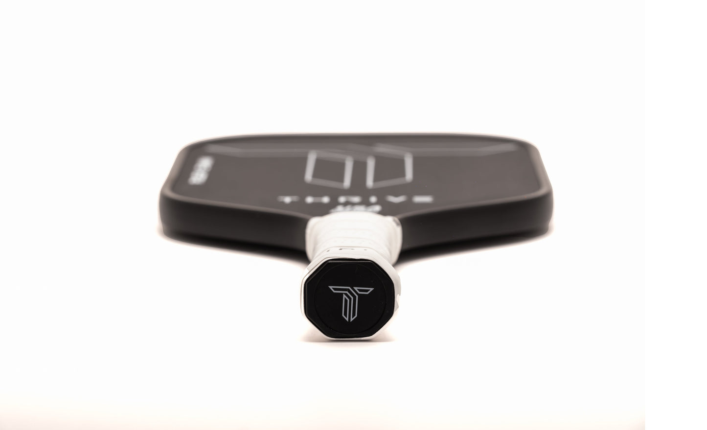 A close-up of the Thrive Pickleball THREAT 16 paddle with a white grip and stylized "T" logo on the handle. The blurred carbon fiber face shows minimalist white lines, “THRIVE,” and includes cover, eraser, custom weight card, and lead weights.