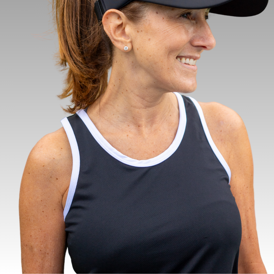 A smiling woman with light brown hair in a ponytail wears the Playly Store Pretty Tank, a black moisture-wicking sports top with white trim, and a black visor. She has stud earrings and stands against a smooth light gray gradient background.