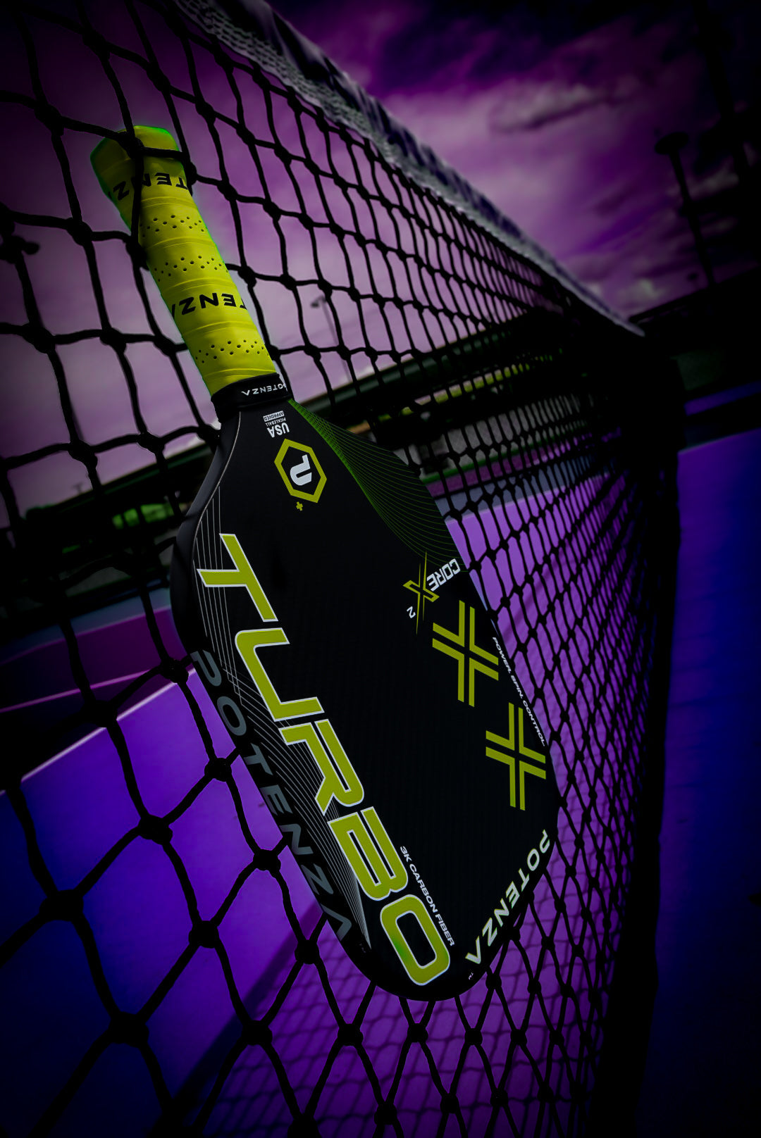 A black and neon yellow Potenza Pickleball TURBO+ COREx2 paddle is wedged in the net on a purple-hued outdoor court, with dramatic lighting highlighting this dynamic carbon fiber pickleball paddle.