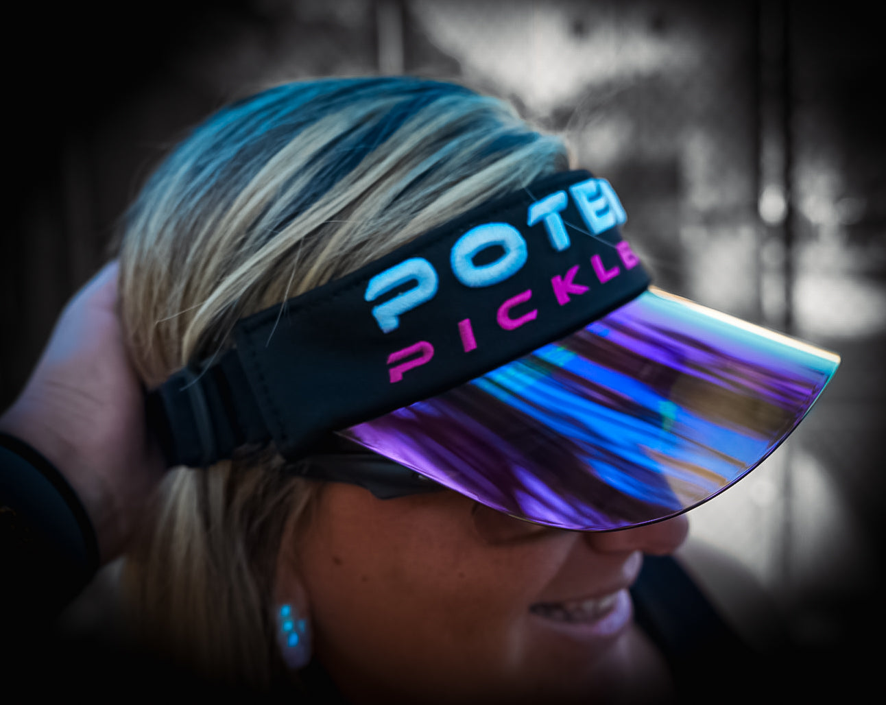 A smiling person with blonde hair wears a Potenza Pickleball Performance Solar Visor with a shiny, holographic brim. The background is blurred, and the cap’s embroidery reads "Potenza Pickleball" as they hold the visor, partially facing the camera.