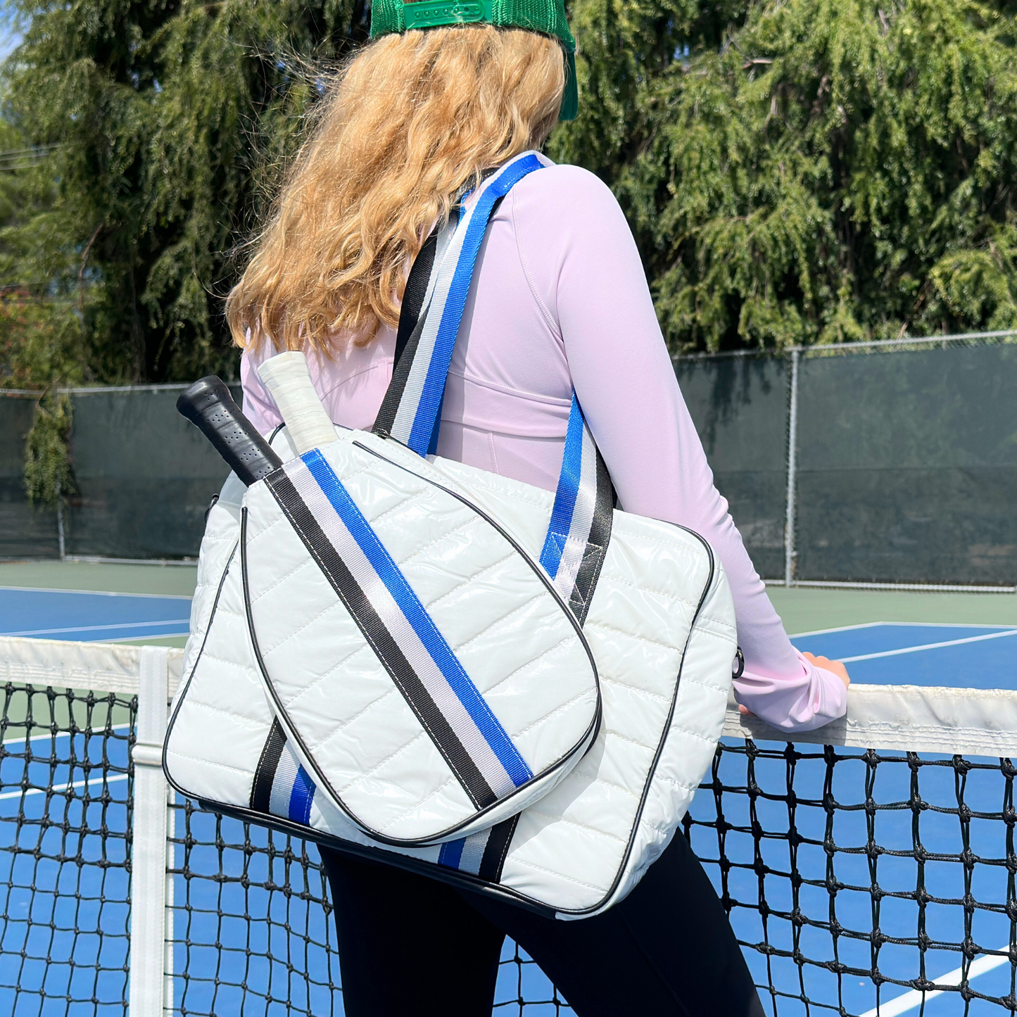 A person with long blonde hair, in a green cap, lavender top, and dark leggings stands by a tennis net carrying the Queen of the Court Kelsey Pickleball Bag—a white quilted ladies bag with blue and black straps. Green trees are in the background.