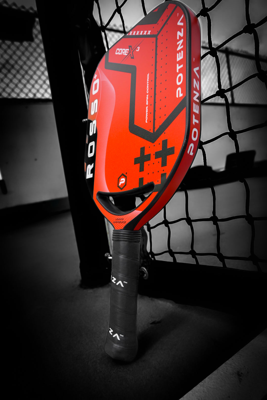 The Potenza Pickleball ROSSO+ COREx3 padel racket, in red with black accents and advanced COREx3 technology, stands upright against a net on the court, its vibrant color and POTENZA branding contrasting a monochrome background.