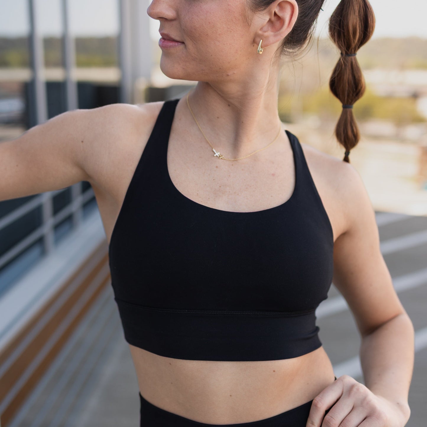 A woman wearing the DYM Athletics Zone Bra (A-D+ cup, High Support) stands outdoors with braided brown hair, looking to the side. Blurred urban buildings and a railing suggest a rooftop women’s activewear setting.