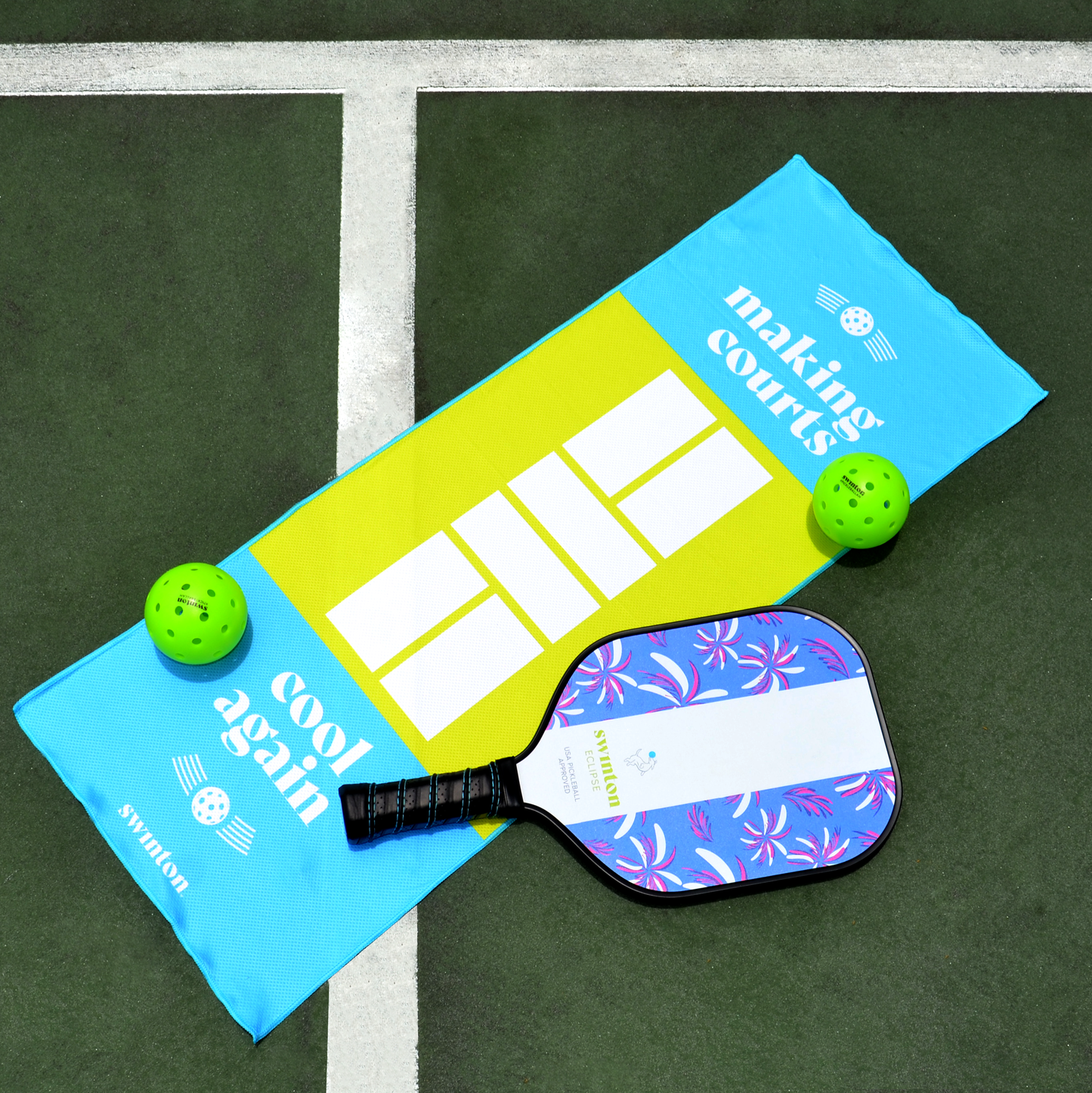 A purple floral pickleball paddle, two green balls, and a Swinton Pickleball Court Cooling Towel reading "making courts cool again" rest on an outdoor court with white lines and a dark green surface.