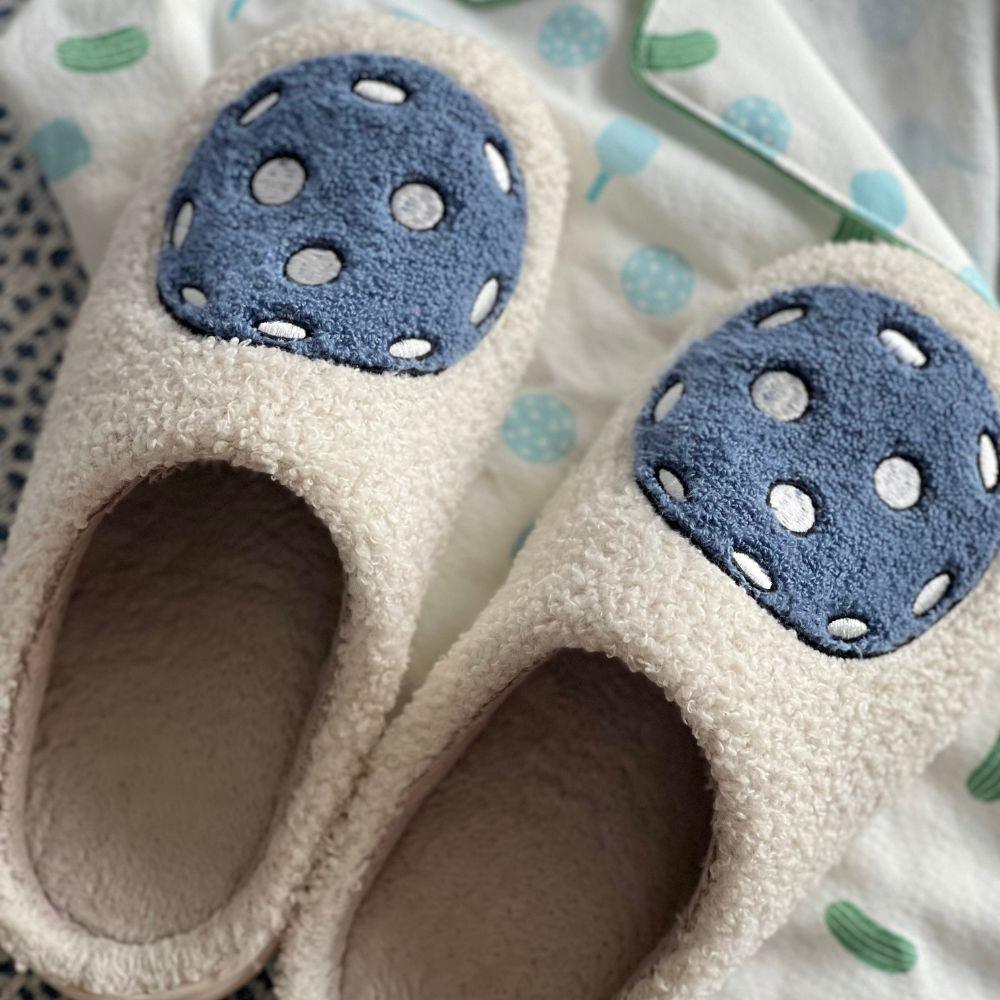 Fresh Pickle's Pickleball Slippers are cozy, cream-colored with a fuzzy texture and blue circular patches with white dots. They rest on soft, white fabric decorated with scattered blue and green shapes.