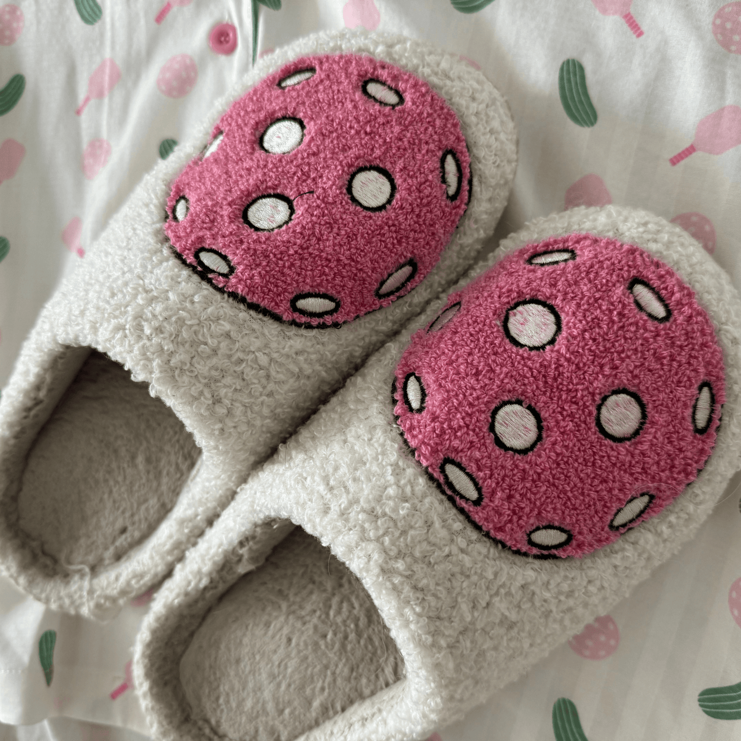 Fresh Pickle’s cozy, cream-colored Pickleball Slippers with soft, fuzzy texture and pink patches featuring white polka dots and black outlines rest on a patterned bedsheet with green, pink, and light red abstract shapes.