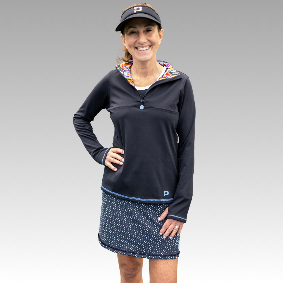 A smiling woman stands against a gray gradient background, wearing the Playly Store Sammy Skort 2.0 in a navy blue pattern, paired with a black CoolFlow zip-up top featuring a colorful inner collar and black visor. One hand rests on her hip.