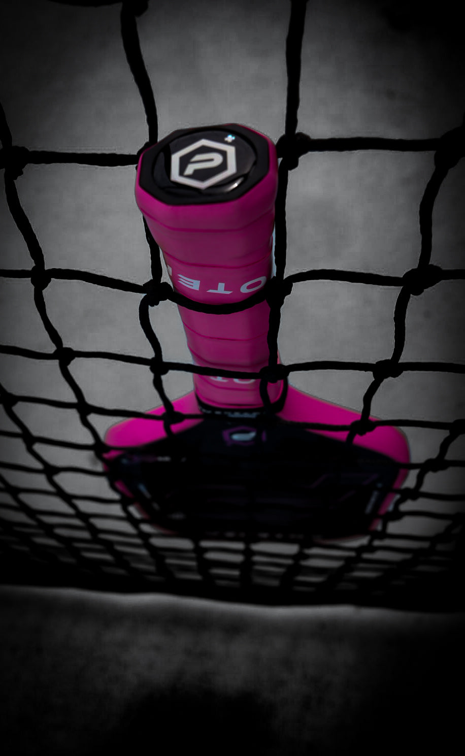 A close-up shows the Potenza Pickleball P+ PowerSpin Carbon COREx3 (Neon Pink) paddle with a vivid pink grip standing upright against a black net. The carbon fiber paddle’s sharp details and neon pink grip stand out against the mostly black-and-white background.