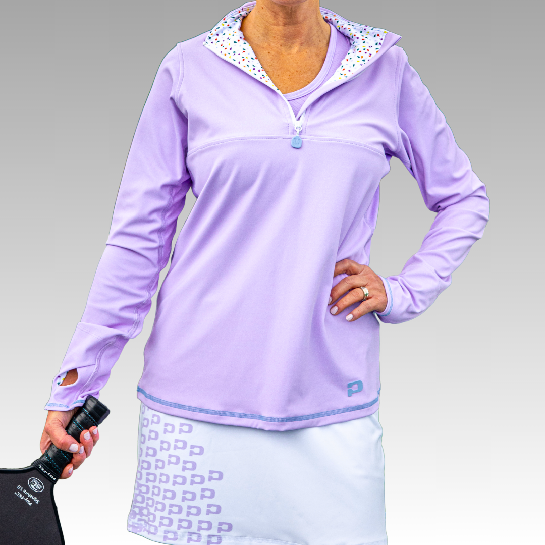 A woman wears the Playly Store Soleil Tech Shirt, a lavender zip-collar top with dotted lining, and a white lavender-pattern skirt, holding a black pickleball paddle against a gray gradient background.