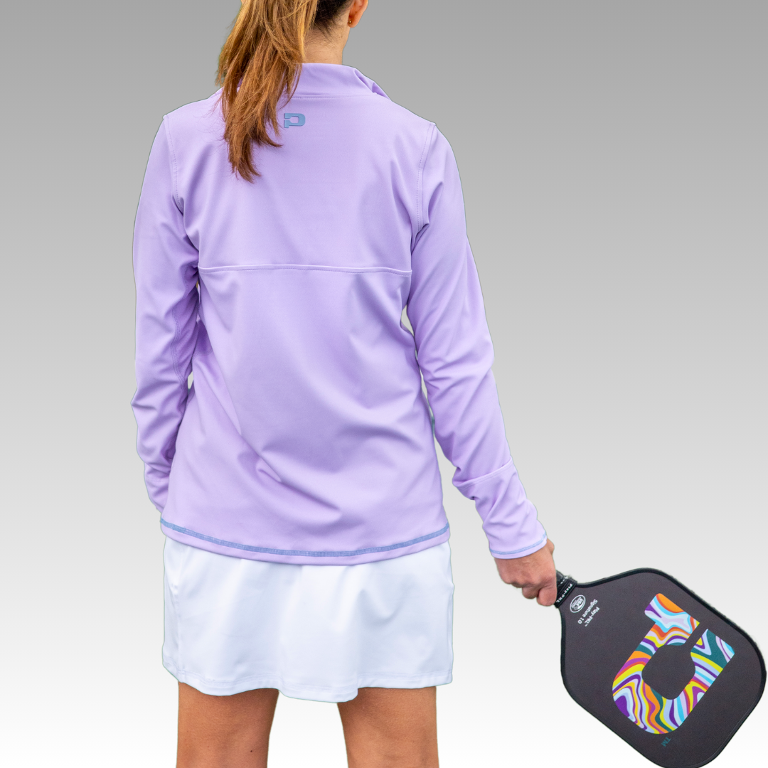 A woman with light brown hair in a ponytail stands facing away, wearing the lavender Playly Store Soleil Tech Shirt and a white skirt. She holds a vibrant, multicolored pickleball paddle against a neutral gradient background.