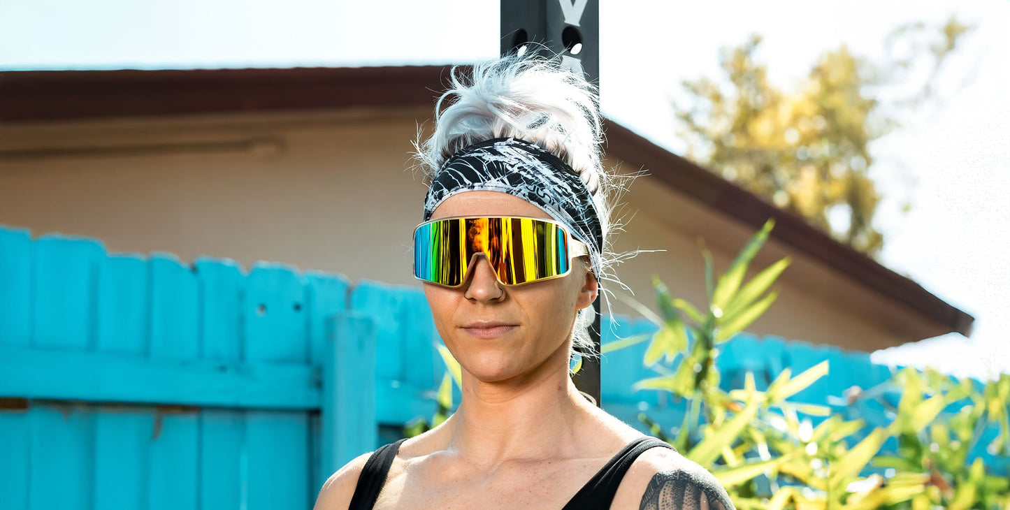 A person with light hair tied back wears Neven Eyewear's Energy shield sunglasses and a black tank top, standing outdoors by a bright blue fence and plants. A tattoo is visible on their arm, complemented by a black-and-white headband.