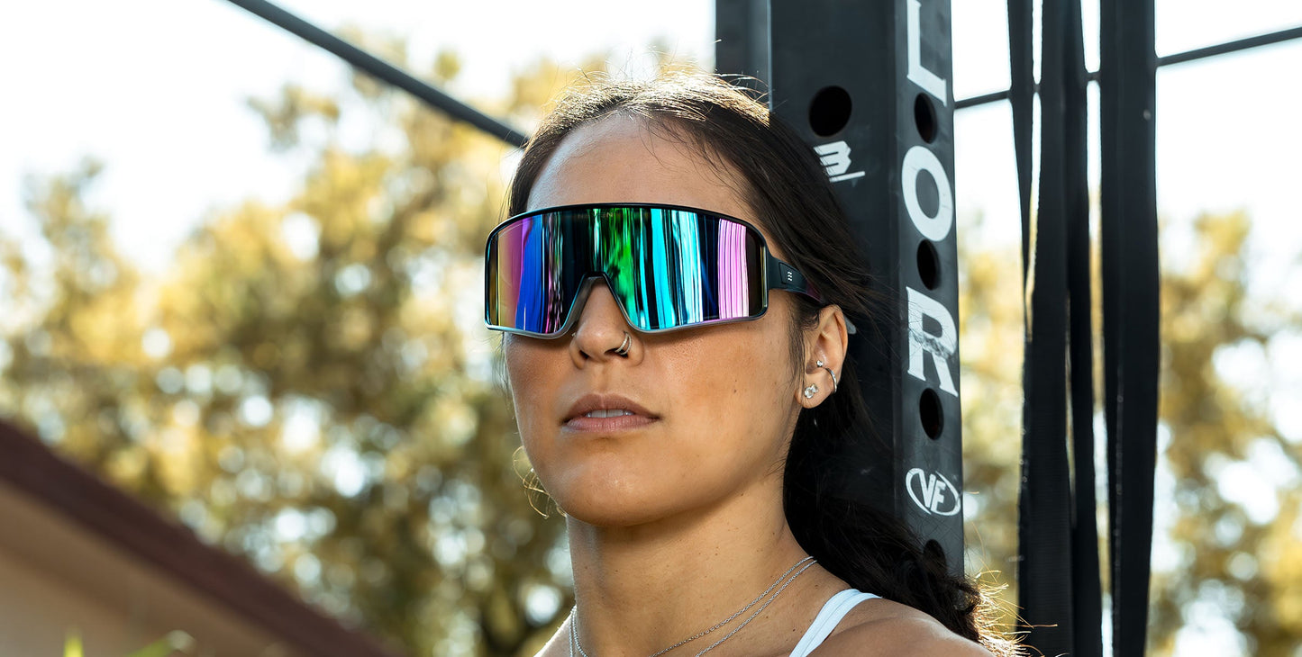 A woman with long dark hair and a nose ring wears oversized Neven Eyewear G.O.A.T sunglasses with polarized UV400 rainbow-mirrored lenses, standing by black exercise equipment in the sun. She also has small earrings.