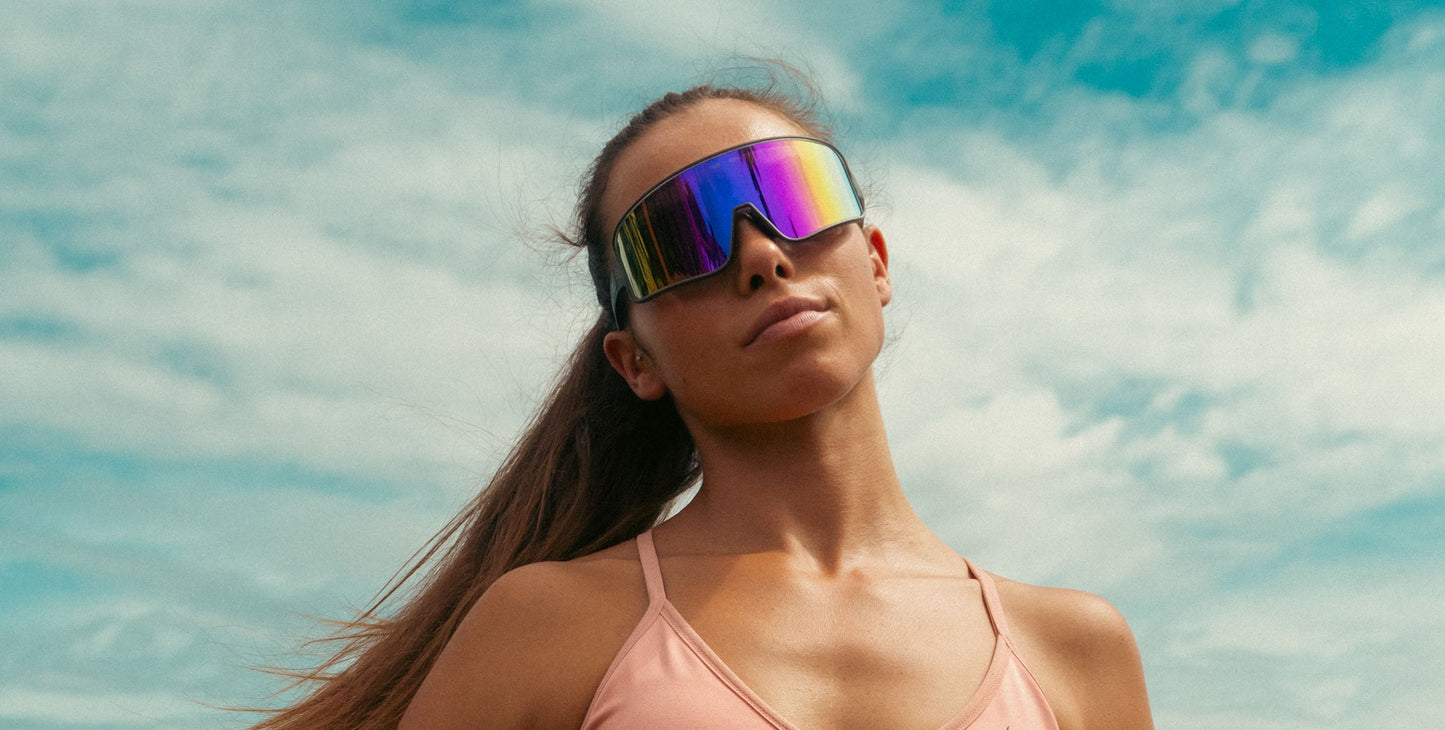 A confident woman with long brown hair in a ponytail models Neven Eyewear's Mattingly oversized visor sunglasses and a light pink sports bra, set against a vintage baseball-inspired backdrop beneath a blue sky.
