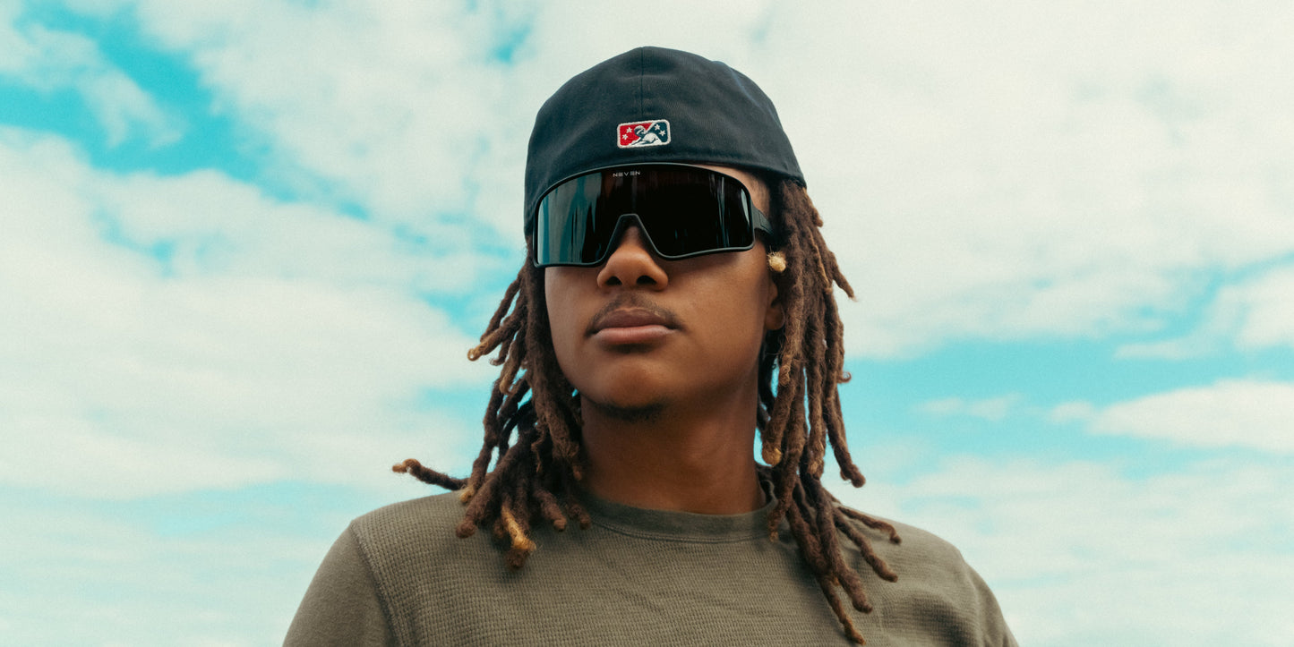 A young person in a backward dark cap wears Neven Eyewear's Resilience black shield sunglasses and a textured olive green shirt, standing against a blue and white partly cloudy sky with a calm expression.