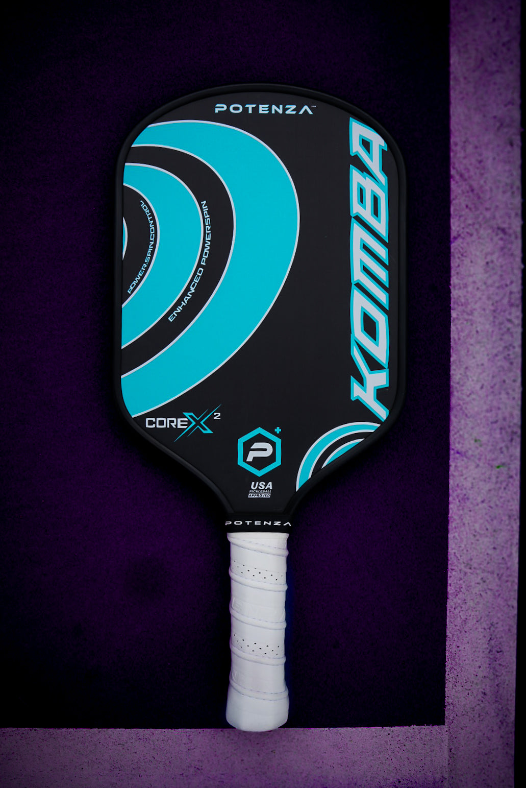 The KOMBA+ COREx2 by Potenza Pickleball is a black carbon fiber paddle with turquoise swirls, "Potenza" and "KOMBA" logos, and “USA Pickleball Approved.” It features a white grip and appears on a purple textured background.