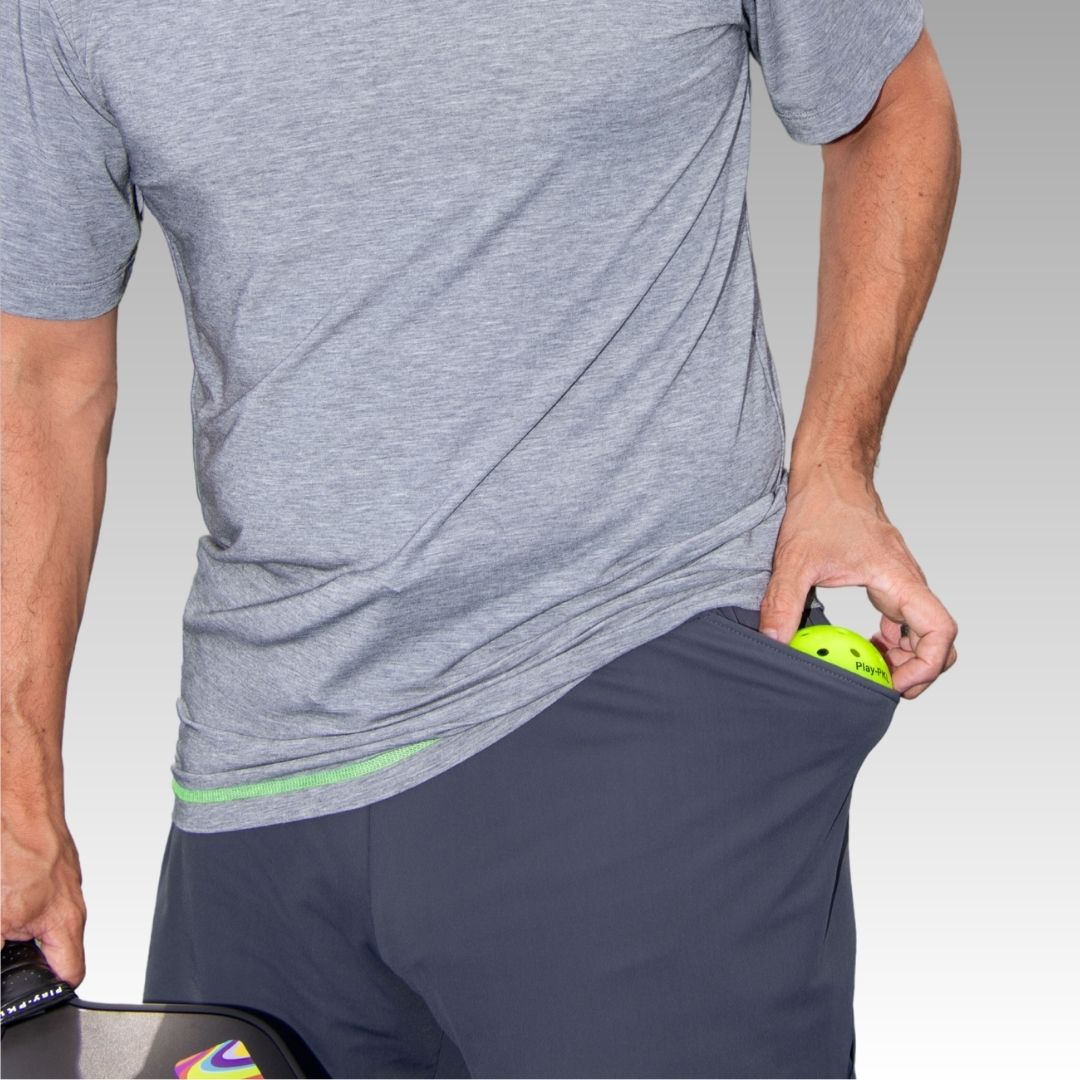 A person wearing Playly Store's Kelter Short and a gray t-shirt places a yellow pickleball into their shorts pocket, holding a paddle in their left hand. The background has a light gray to white gradient. The person's face is not visible.