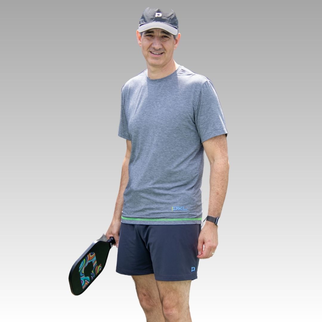 A man stands facing the camera in a gray athletic shirt, black cap, and Playly Store Kelter Short, holding a pickleball paddle by his side. He wears a watch and smiles slightly against a simple gray gradient background.
