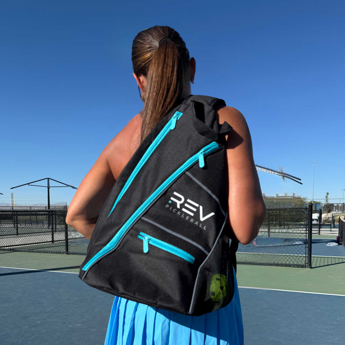 A woman with brown hair in a ponytail stands on an outdoor pickleball court, wearing a blue skirt and carrying the REV Pickleball ELITE Paddle Bag with turquoise zippers over her right shoulder under a clear blue sky.