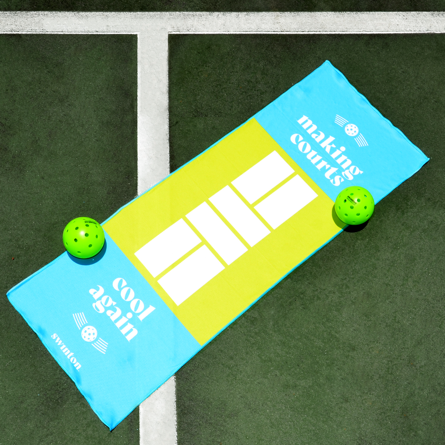 The Swinton Pickleball Court Cooling Towel, featuring blue and yellow colors, a court graphic, and "making courts cool again," rests on an outdoor pickleball court beside green perforated balls and crisp white lines.