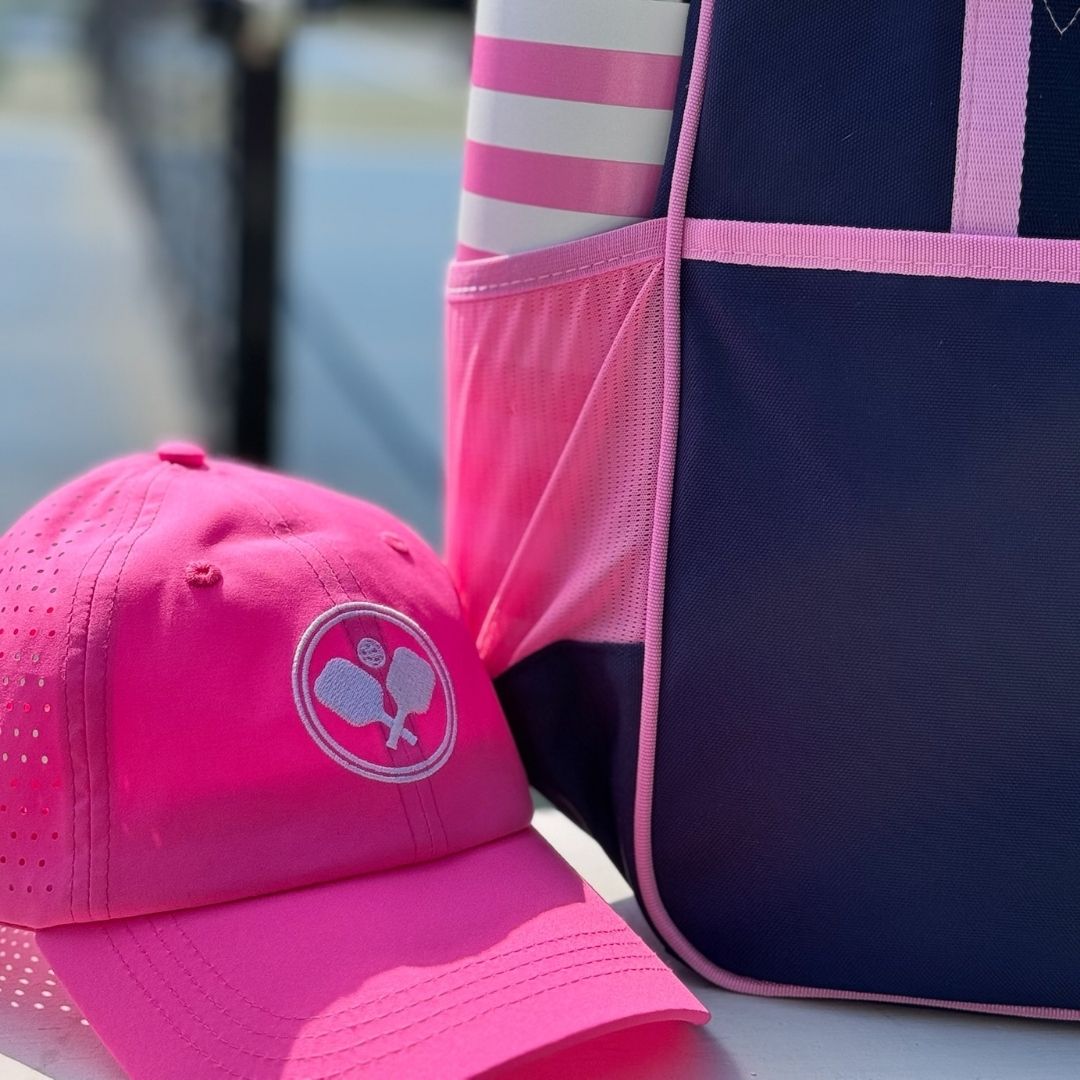 A Fresh Pickle Pickleball Cap in vibrant pink, featuring a tennis ball and crossed racket logo, rests next to a navy and pink sports bag with mesh and stripe details. Sunlight adds an athletic vibe while the background stays blurred.