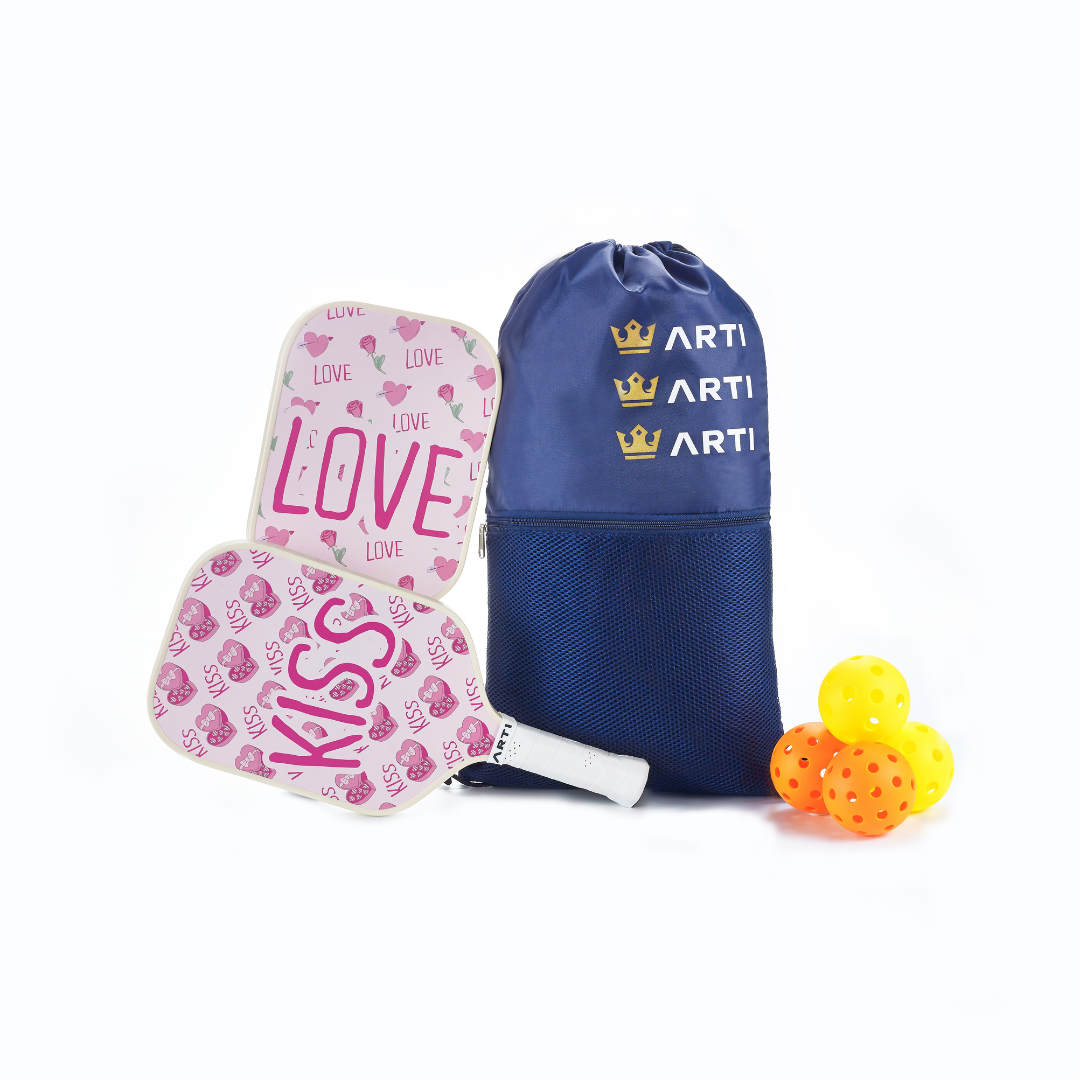 The ARTI Fiberglass "Valentine's" Pickleball Paddle Set features two pink and white heart-themed paddles, three yellow pickleballs, and a blue ARTI drawstring bag with gold crowns, all displayed on a white background.