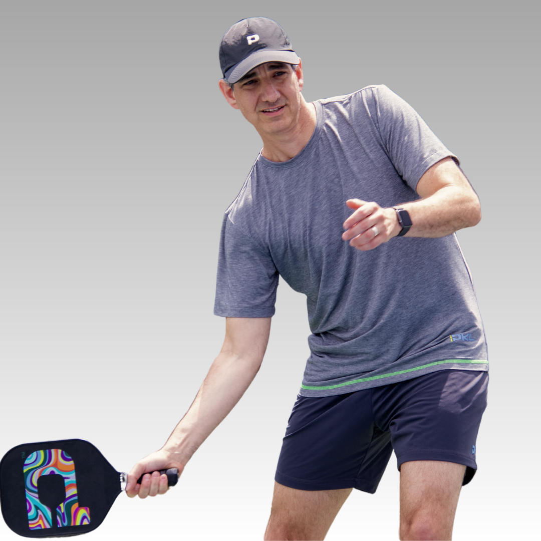 A man in an athletic fit Playly Store Warrior Tee, navy shorts, and a cap gets ready to hit a ball with a colorful paddle. Focused and in mid-action, he extends his right arm against a plain gray gradient background.