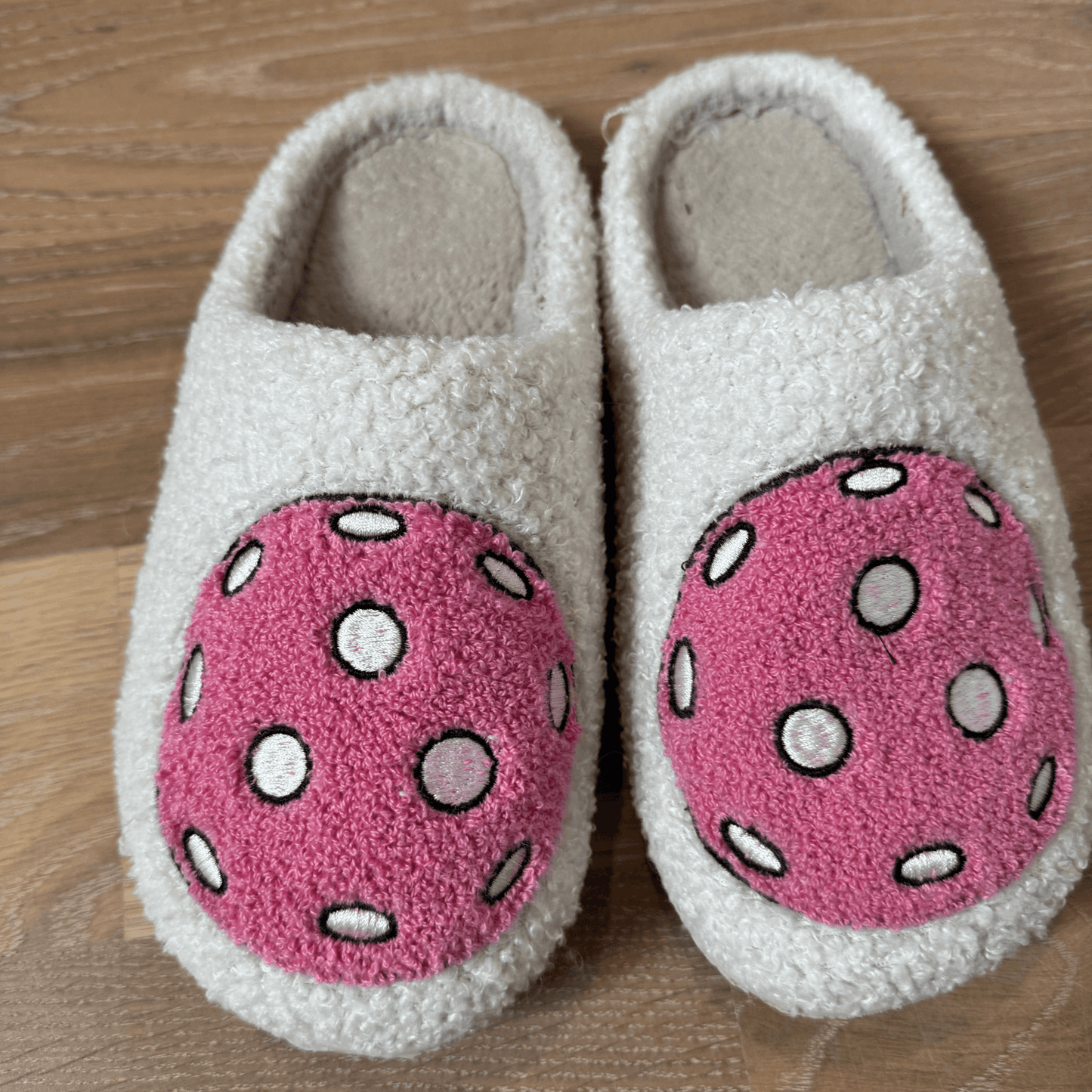 Fresh Pickle's Pickleball Slippers are fuzzy and white with large pink circles featuring white and black polka dots on the front. The comfy slippers are shown side by side on a wooden floor.
