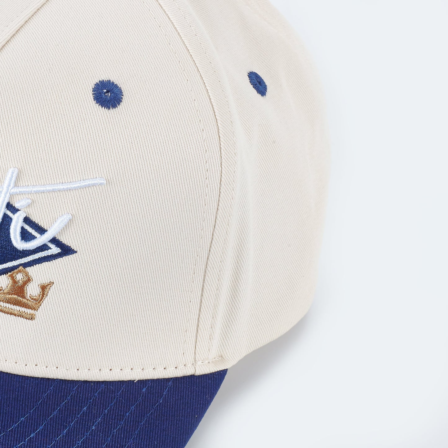 Close-up of the ARTI Throwback Classic Rope Hat by ARTI in beige and blue, featuring blue ventilation holes, a blue brim, white and blue side embroidery, and a small gold crown design against a light gray background.