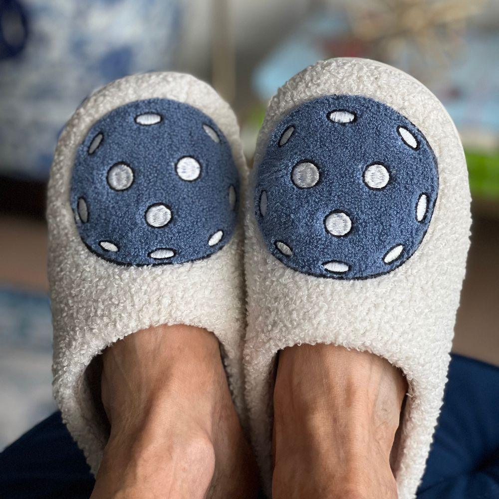 A close-up of two feet wearing Fresh Pickle Pickleball Slippers. Each fuzzy white slipper has a blue circular patch with white polka dots, set against a blurred background that highlights the cozy Fresh Pickle footwear.