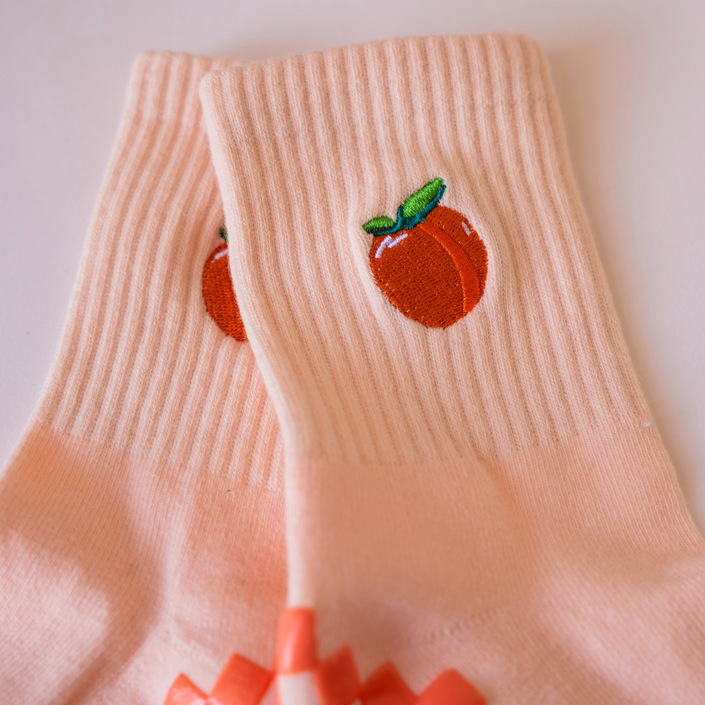 The Just Peachy Grip Sock by Revel features pale pink ribbed fabric with embroidered orange peaches and green leaves near the ankle. Overlapping on a white surface, these socks are a playful addition to your Pilates collection.