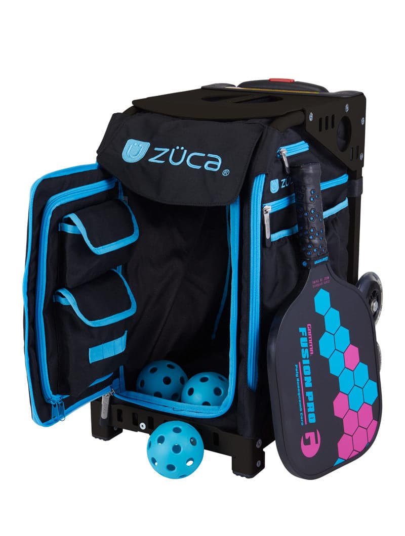 The ZUCA Pickleball Sport Black/Blue bag stands open to reveal three yellow pickleballs inside. It features multiple pockets, a zippered flap, and a black paddle with blue and pink accents attached to the side. Two wheels are visible at the back.
