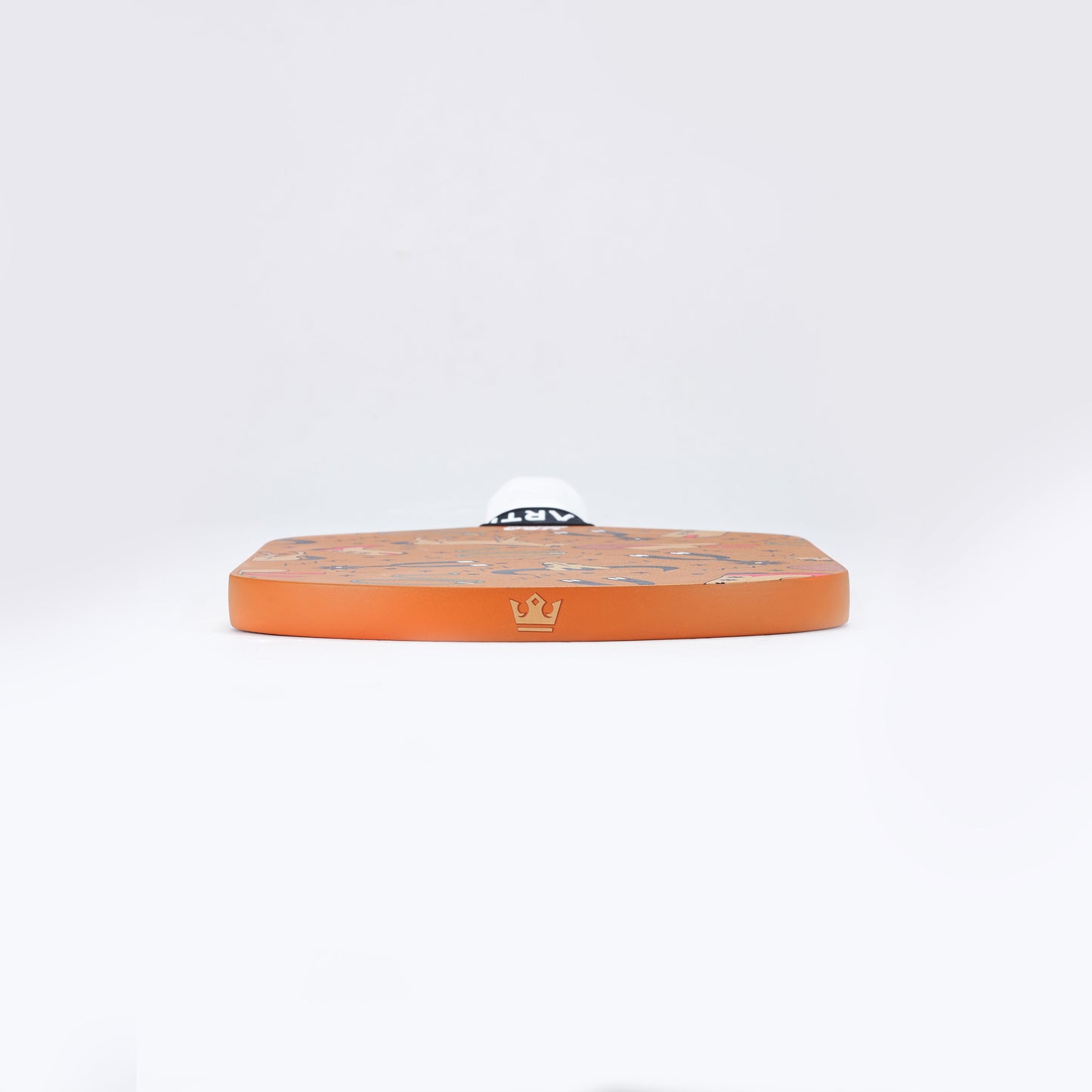 The ARTI Carbon Fiber "TEXAS" Paddle, featuring a flat orange hexagonal design with a gold crown emblem, is displayed on a white background. A small white ball with black text sits partially visible at the back edge.