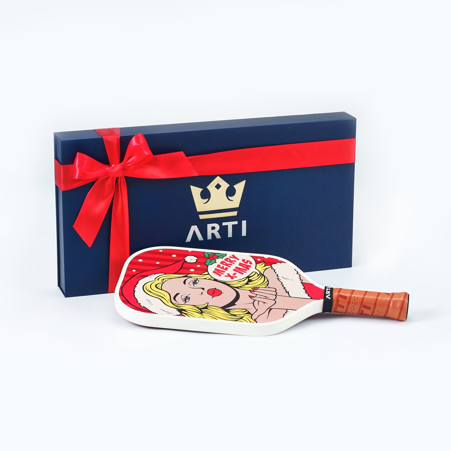 A blue ARTI gift box with a red ribbon and gold crown logo stands behind the ARTI Carbon Fiber Kristen&Kristy "X-MAS" Paddle, a USAPA-approved Christmas paddle with pop art of a blonde woman in a Santa hat and a tan grip handle.