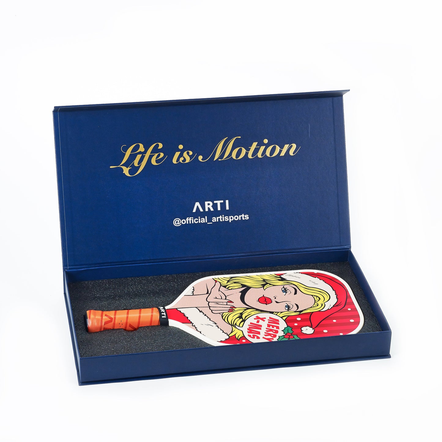 A blue box with gold "Life is Motion" text holds the ARTI Carbon Fiber Kristen&Kristy "X-MAS" Paddle, USAPA approved and featuring a pop art blonde woman in a Santa hat. The handle is orange and ARTi branding appears inside the box.