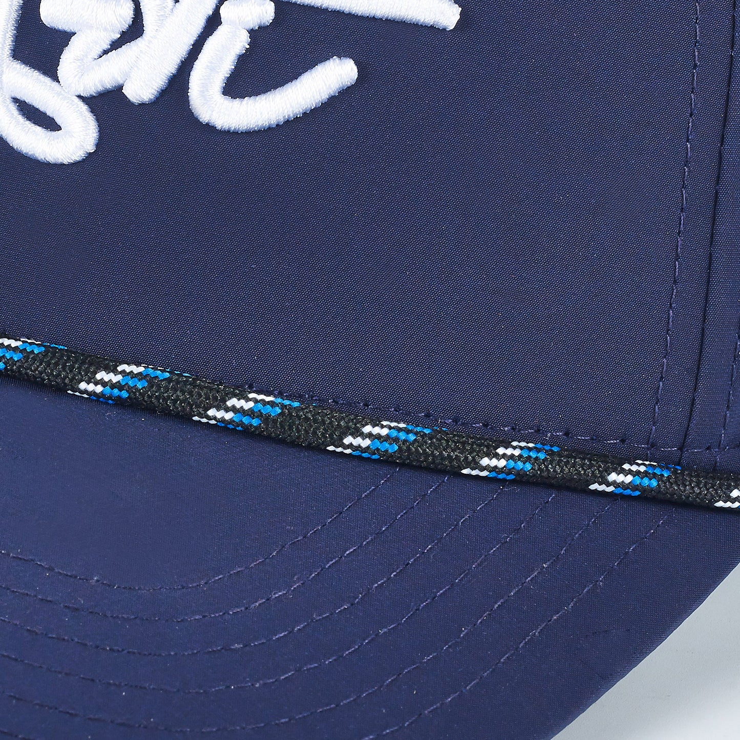 Close-up of the ARTI Navy Performance Rope Mesh Classic Hat by ARTI, featuring white script embroidery and a black, white, blue, and green braided cord above the stitched brim for a timeless look.
