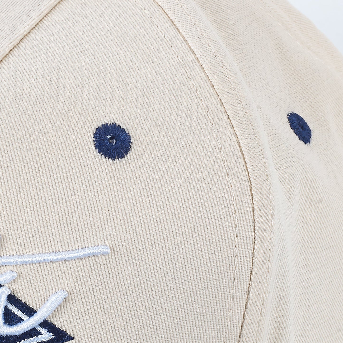 Close-up of the ARTI Throwback Classic Rope Hat by ARTI in beige, featuring two navy blue embroidered eyelets and part of a raised white and navy logo on the front. The hat’s texture and stitching details are prominently visible.