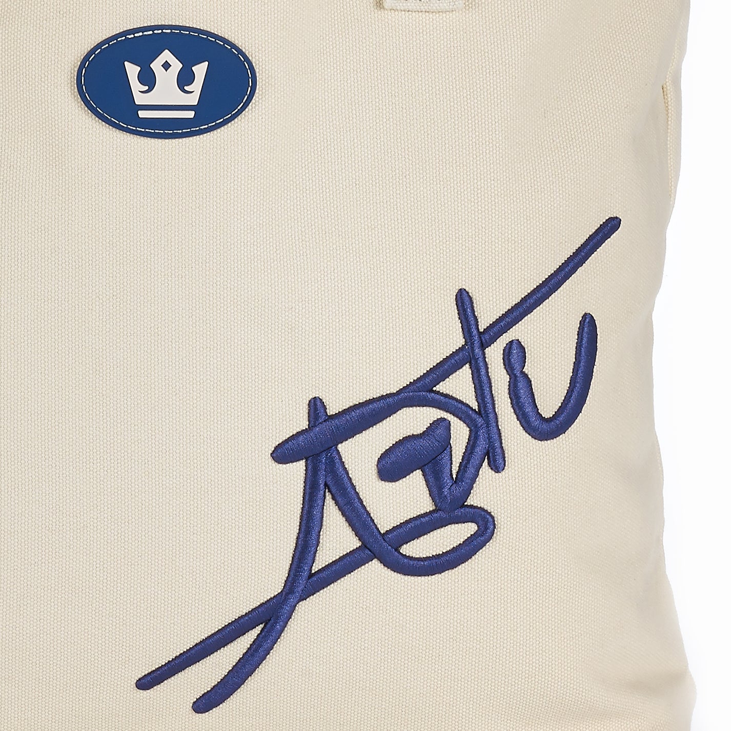 A close-up of the ARTI Cream Tote Bag by ARTI, made from cotton with a blue embroidered signature and a round blue patch featuring a white crown. The detailed stitching and fabric texture showcase this eco-friendly tote's quality.