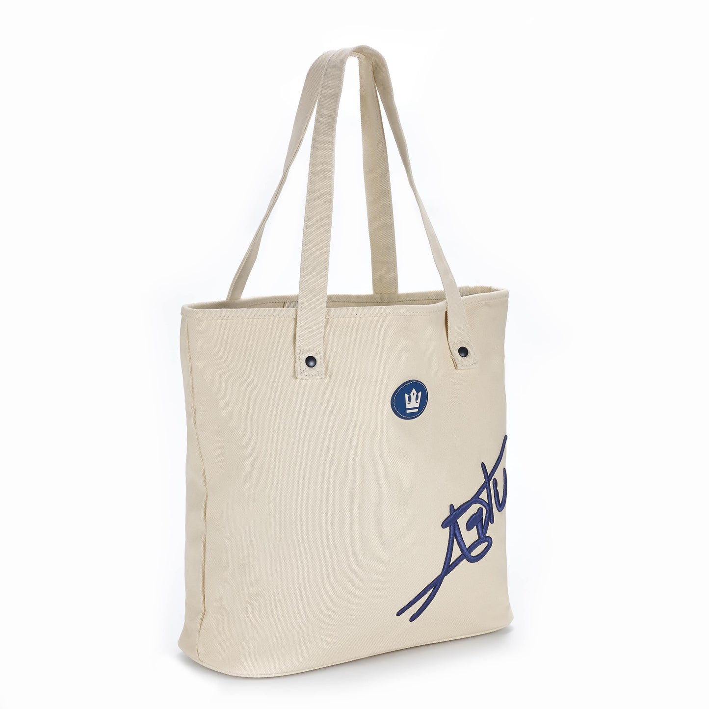 The ARTI Cream Tote Bag by ARTI is a cotton tote with two handles, silver rivet details, and a blue circular patch featuring a white crown and bold blue script on the front. This eco-friendly bag is shown against a plain white background.