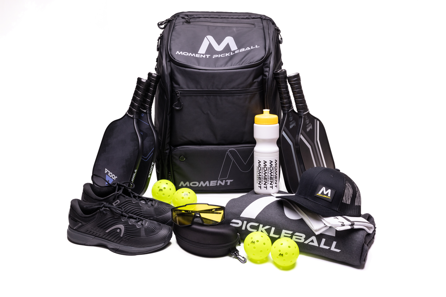 The Moment Pickleball MW-PRO Bag / Travel Bag in Black/Silver is placed among four black paddles, three yellow pickleballs, shoes, a white water bottle, sunglasses, a cap, and a gray towel—all arranged against a white background.