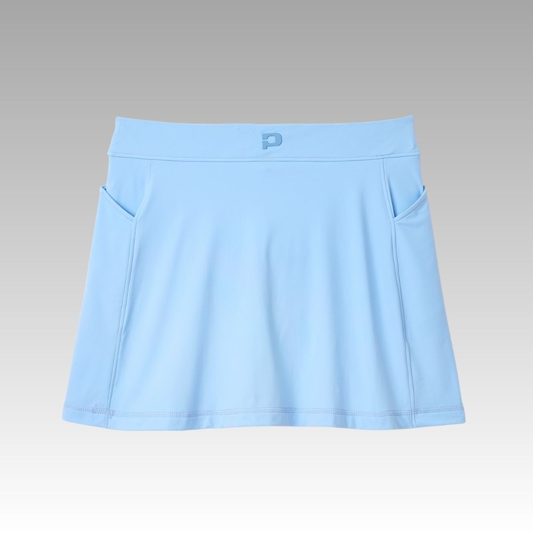 The Playly Store Sammy Skort is a light blue athletic skort with a wide waistband, two side pockets, and a subtle logo. Made from SofKnit moisture-wicking fabric, it's shown against a smooth gray-to-white gradient background.