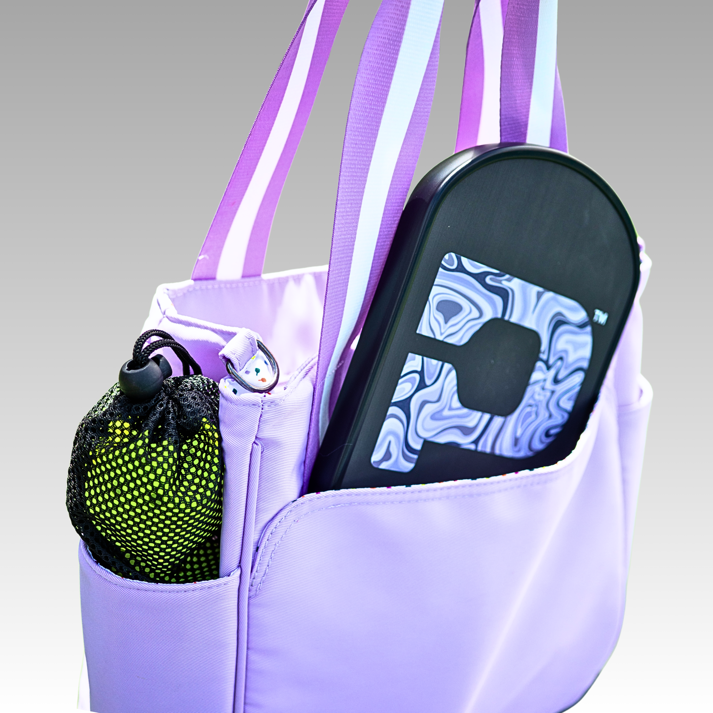 The Lulu Pickleball Tote by Playly Store features striped handles, holds a black-and-white patterned paddle, and has an outer pocket with a mesh bag containing two yellow-green pickleballs, all set against a gradient gray background.