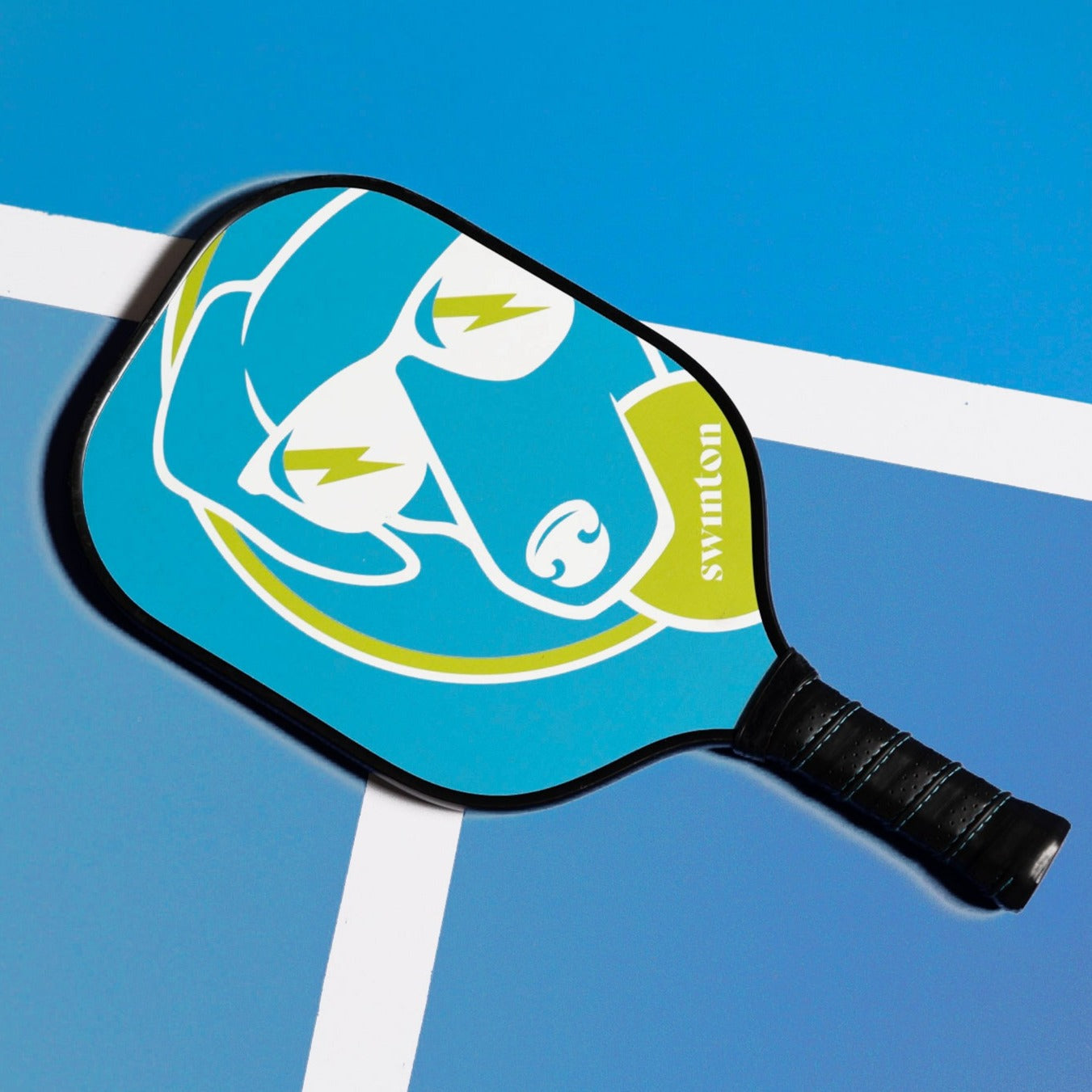 The Swinton Pickleball Hero Blue Dog Pickleball Paddle is a lightweight graphite paddle with a black handle, blue face, and features a sunglasses-wearing dog and yellow lightning bolts on court, plus "swinton" in white.